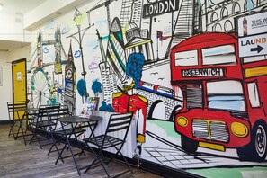 A room with a mural of London landmarks, a red double-decker bus labeled 'Greenwich', and foldable chairs. - St Christopher's Inn, Greenwich - Hostel (London)