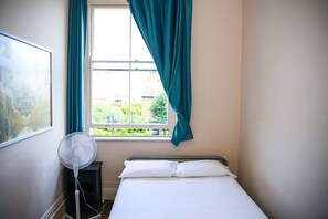 A room with a bed, a window with curtains, a standing fan, and a painting on the wall. - St Christopher's Inn, Greenwich - Hostel (London)