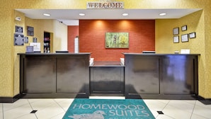A reception area with a 'WELCOME' sign, a reception desk, a 'HOMEWOOD SUITES' mat, and informational posters on the walls.