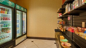 A hotel lobby with a self-service beverage cooler stocked with various drinks, a snack bar with packaged foods, and a clean, tiled floor.
