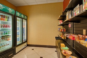 A hotel lobby with a self-service beverage cooler stocked with various drinks, a snack bar with packaged foods, and a clean, tiled floor. - Homewood Suites by Hilton Tulsa-South (Broken Arrow)