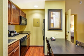 A compact kitchen with wooden cabinets, a microwave, and a coffee maker. There is a dining table with chairs and a framed artwork on the wall.