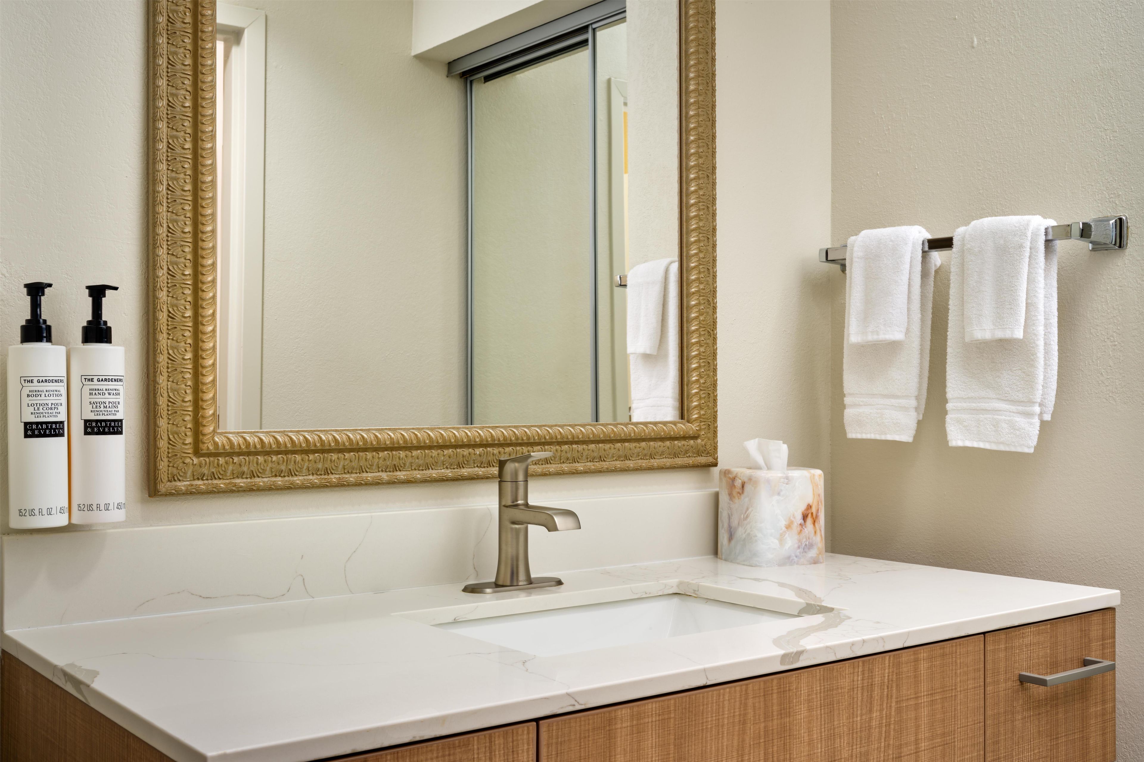 A bathroom with a large mirror, a sink, two bottles of hand wash, a marble soap dish, and a towel rack with towels.