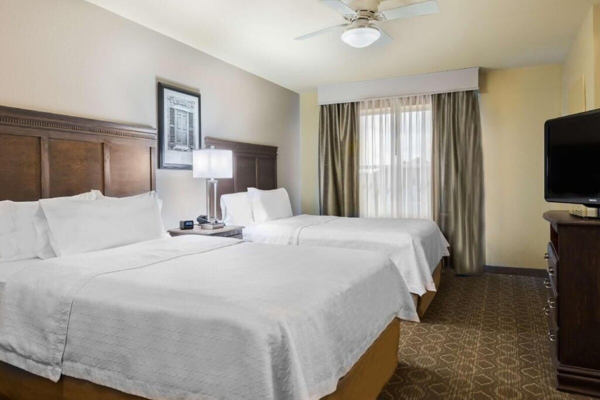A hotel room with two beds, a ceiling fan, a TV, a framed picture on the wall, and a window with curtains.