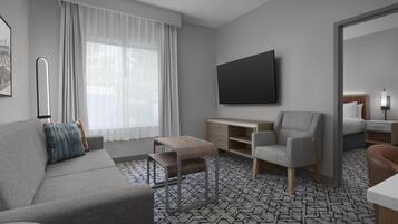 A modern hotel room with a grey sofa, a small wooden desk, a grey armchair, a flat-screen TV, and a patterned carpet.