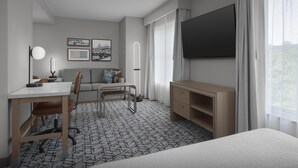 Habitación de hotel con cama, escritorio, silla, sofá, televisor y ventana con cortinas.