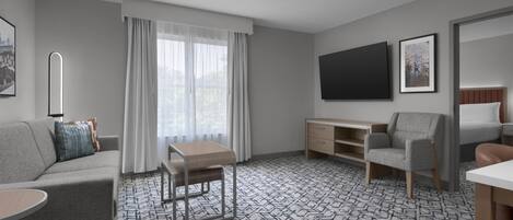 Una habitación de hotel moderna con un sofá gris, un escritorio pequeño, una silla, un televisor y una alfombra estampada.