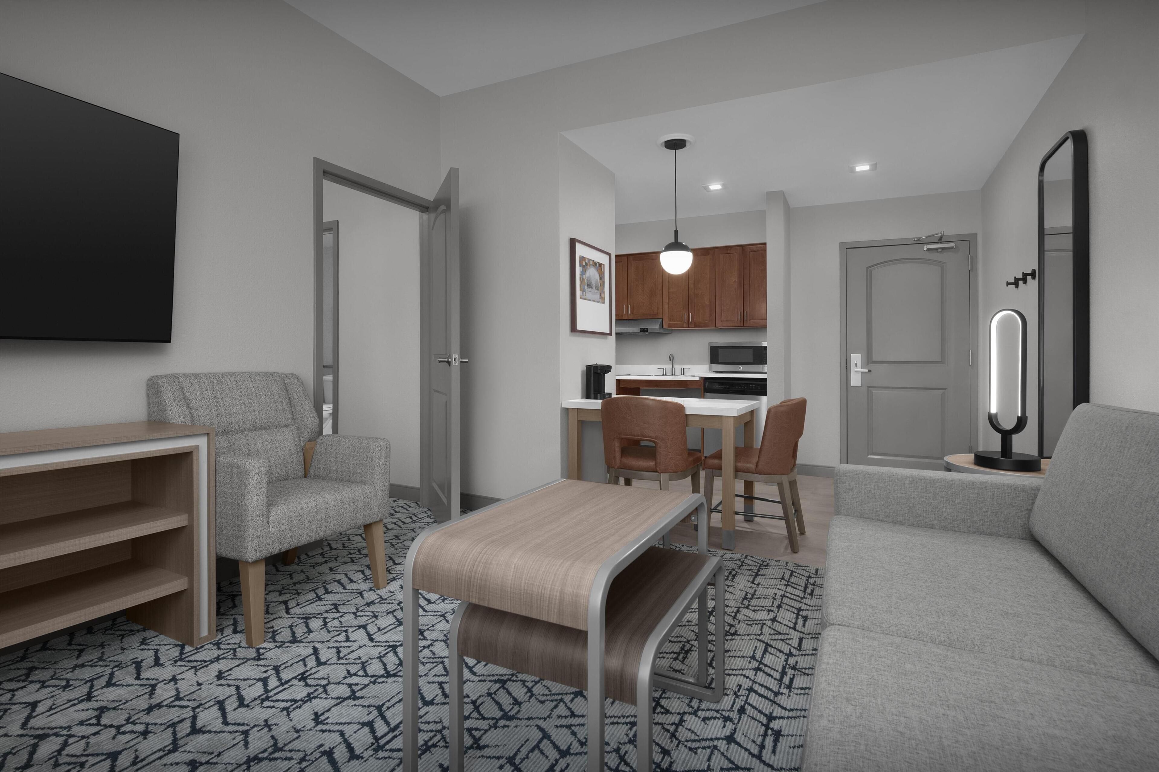Una sala de estar moderna con un sofá gris, una mesa de centro de madera y un tapete estampado. Hay un comedor con mesa y sillas, y una cocina con armarios de madera y un microondas.