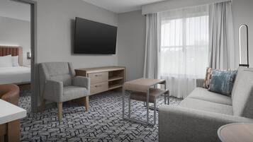 Una habitación de hotel moderna con una cama, un sillón gris, una mesita, un televisor montado en la pared y un ventanal con cortinas traslúcidas.