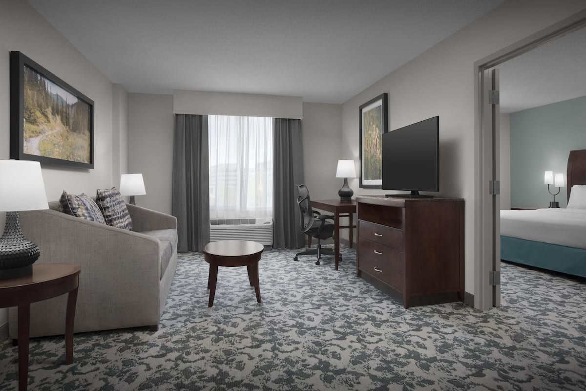 Una habitación de hotel con sofá, escritorio, televisor, cama y ventana con cortinas.