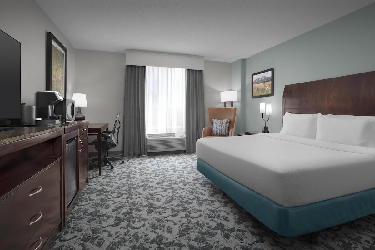 Habitación de hotel con una cama grande, un escritorio con lámpara, una silla y una ventana con cortinas.