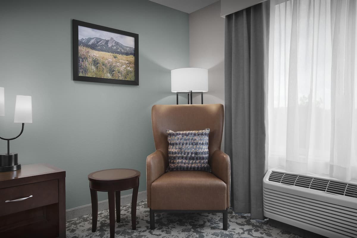 Habitación de hotel con un sillón marrón, una mesita redonda pequeña, una lámpara, un cuadro enmarcado en la pared y una ventana con cortinas traslúcidas.