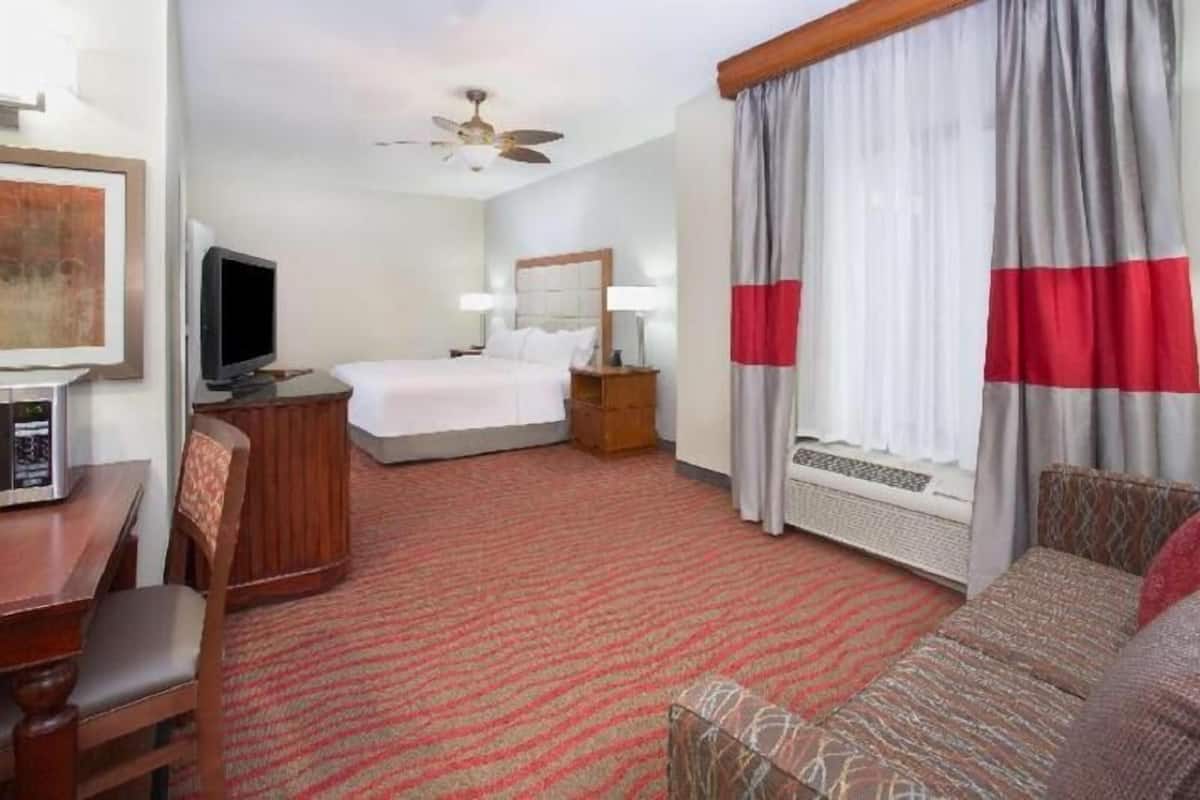 A hotel room with a bed, a desk with a television, a chair, a sofa, a ceiling fan, and curtains.