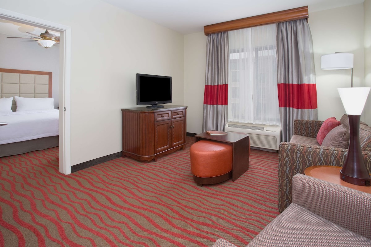 A hotel room with a bed, a TV, a wooden cabinet, a sofa, an ottoman, and a lamp.