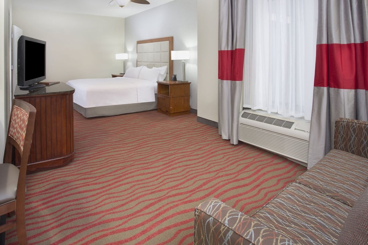 A hotel room with a bed, a desk with a television, a chair, a sofa, a ceiling fan, and a window with curtains.