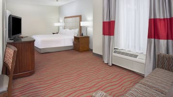 A hotel room with a bed, a desk with a television, a chair, a sofa, a ceiling fan, and a window with curtains.