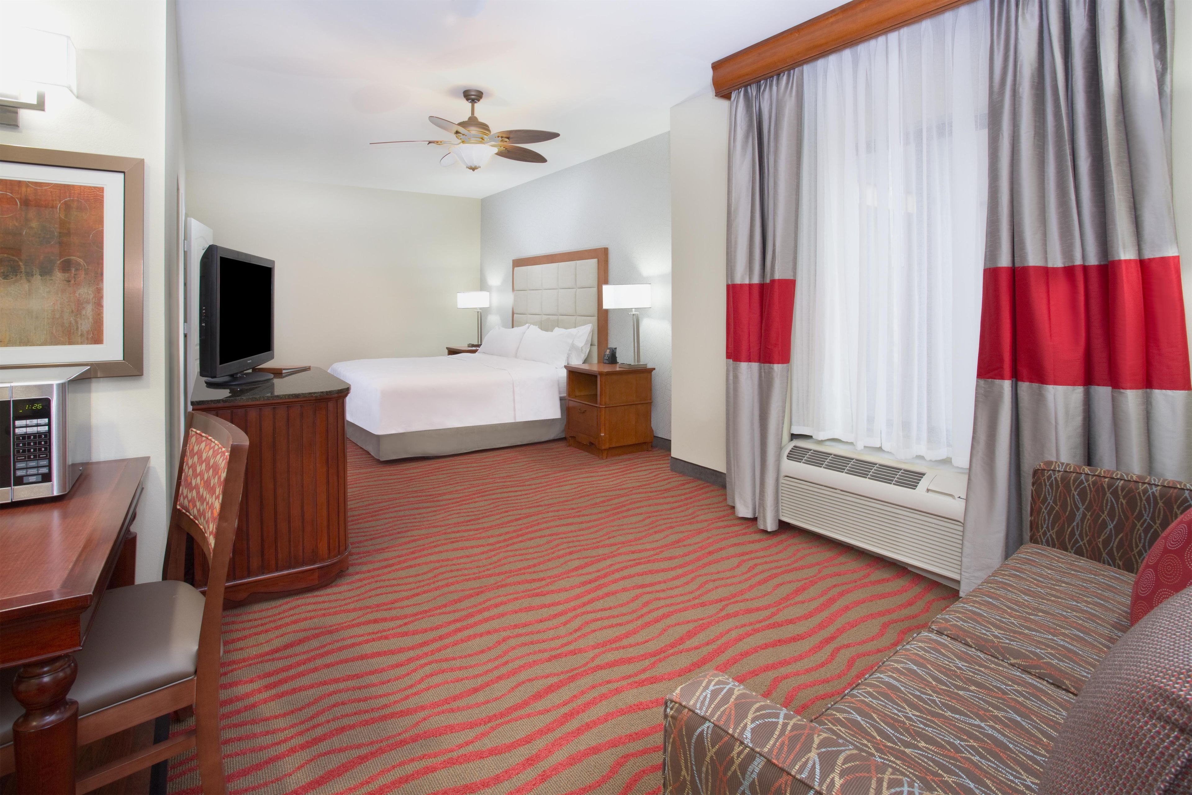 Een hotelkamer met een bed, een bureau, een televisie, een plafondventilator en een raam met gordijnen.