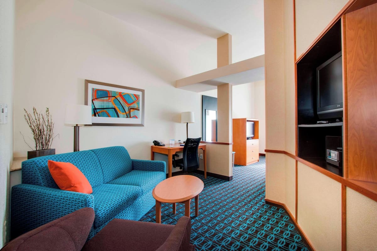 A hotel room with a blue patterned sofa, a small round table, a television, a desk with a chair, and a decorative wall piece.