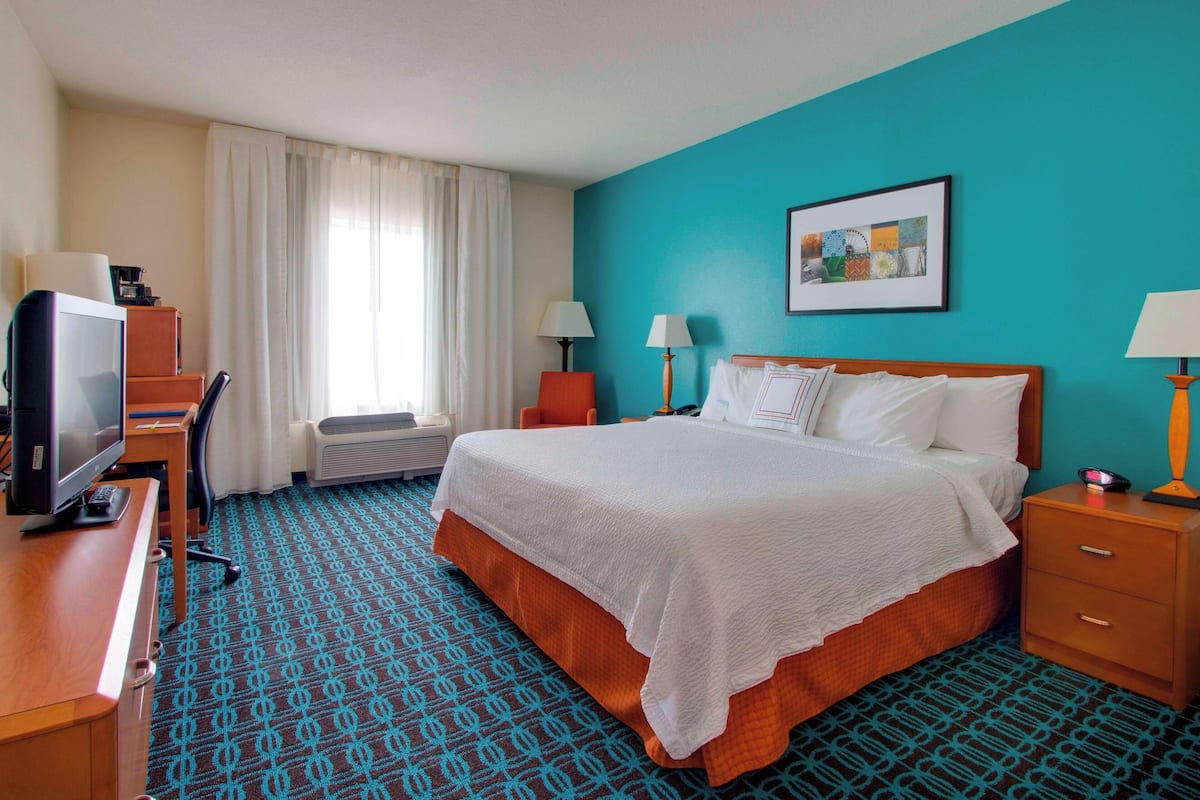 A hotel room with a bed, a desk with a computer, two bedside lamps, a window with curtains, and a wall-mounted artwork.