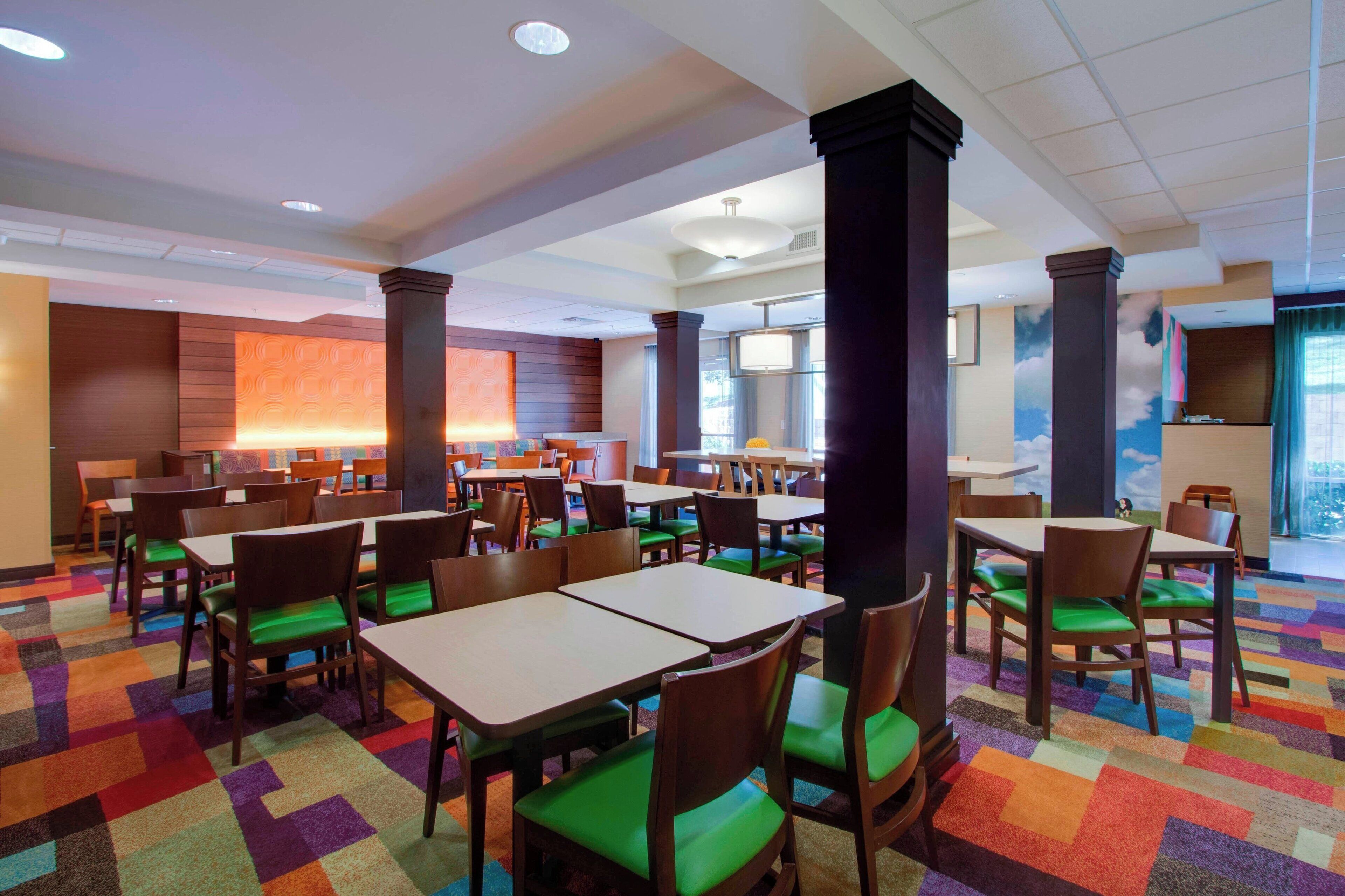 Uma sala de jantar de hotel com várias mesas e cadeiras, um tapete colorido e uma janela grande com vista.
