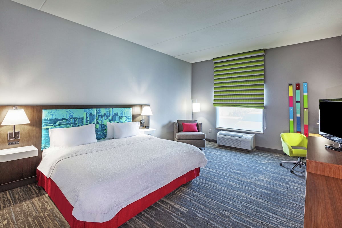 A hotel room with a large bed, a desk, a chair, a TV, a window with blinds, and a cityscape painting on the wall.