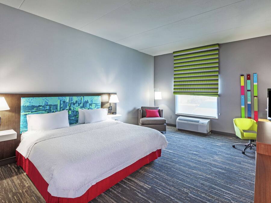 A hotel room with a large bed, a desk, a chair, a TV, a window with blinds, and a cityscape painting on the wall.