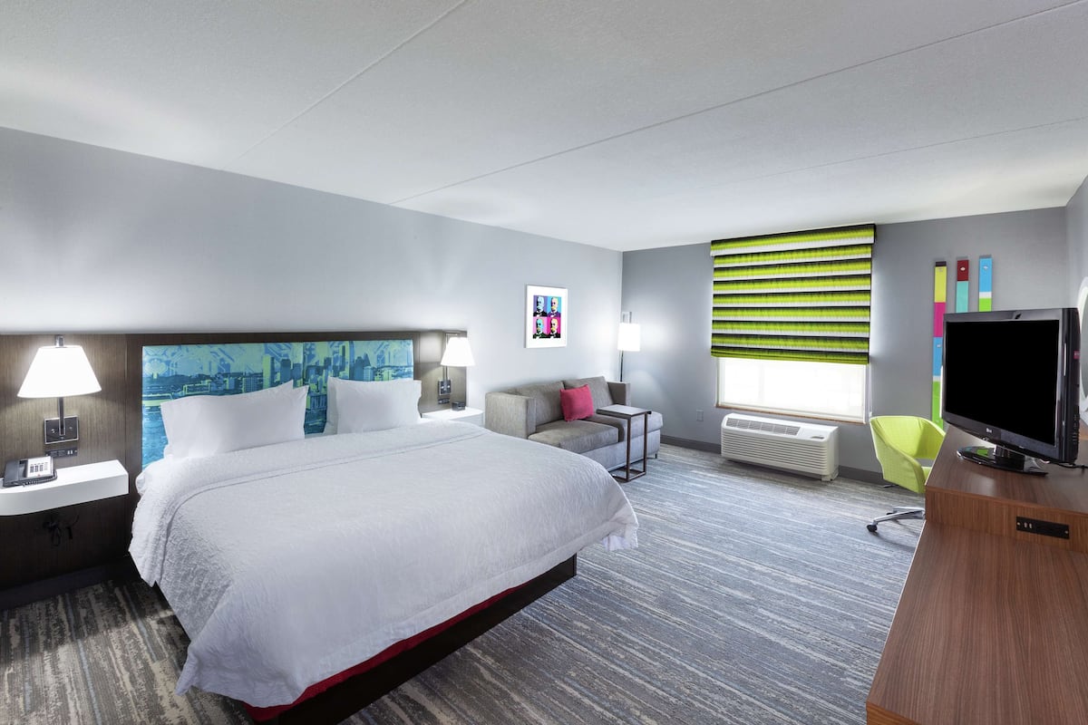 A hotel room with a large bed, a desk with a TV, a chair, a window with blinds, and a wall-mounted air conditioner.