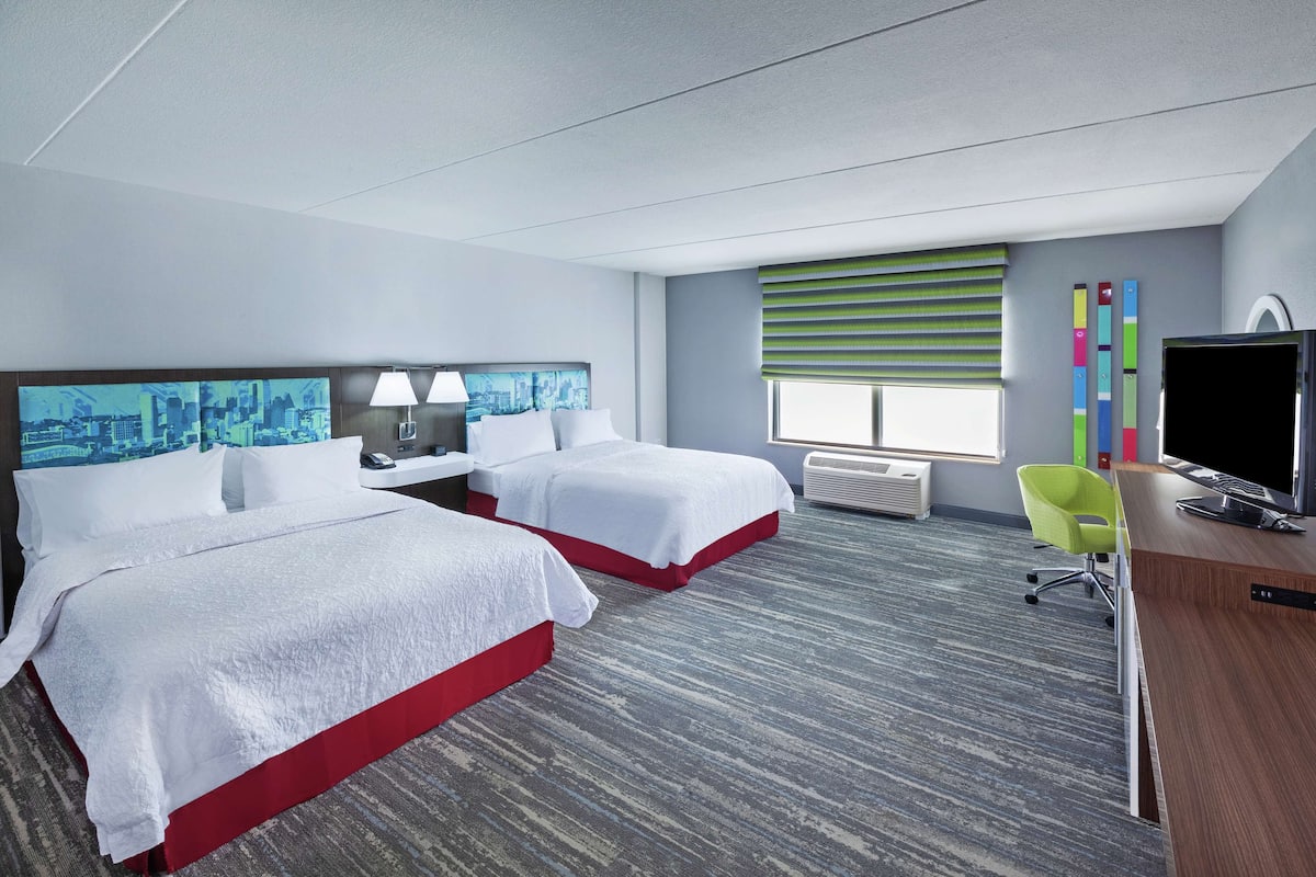 A hotel room with two beds, a desk, a chair, a TV, and a window with blinds.