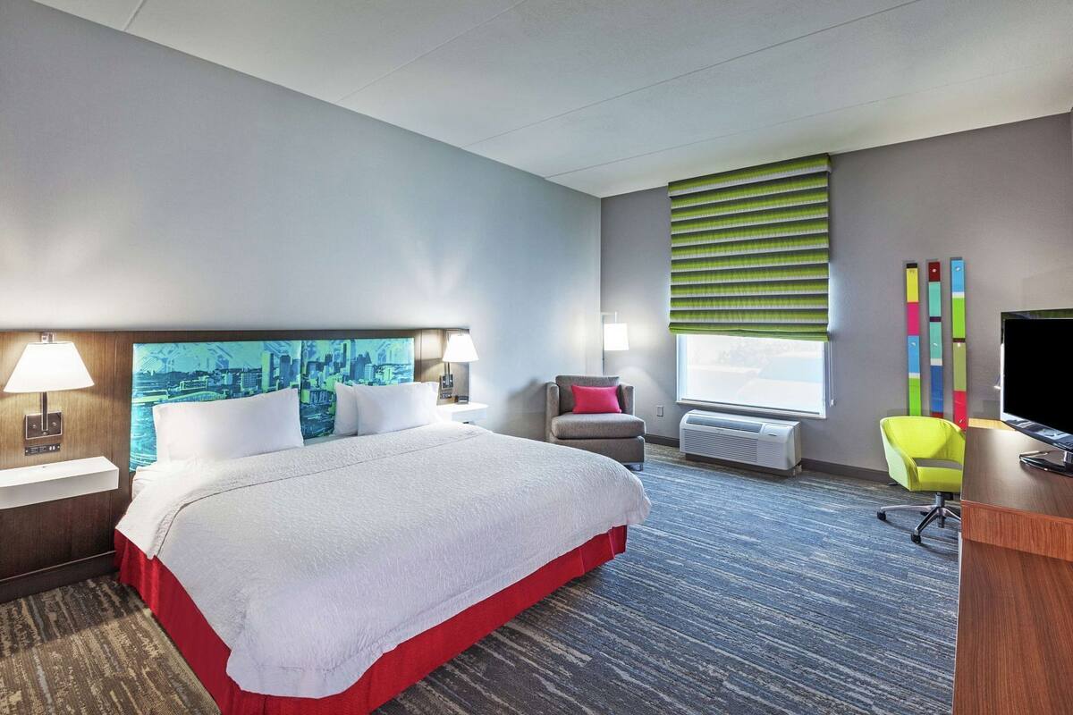A hotel room with a large bed, a desk, a chair, a TV, a window with blinds, and a cityscape painting on the wall.