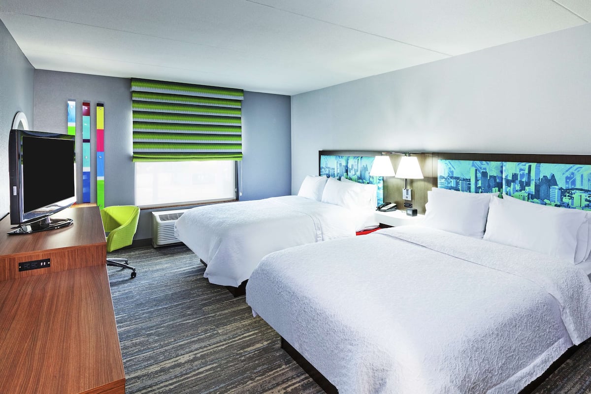 A hotel room with two beds, a desk with a television, a cityscape mural, and a window with blinds.