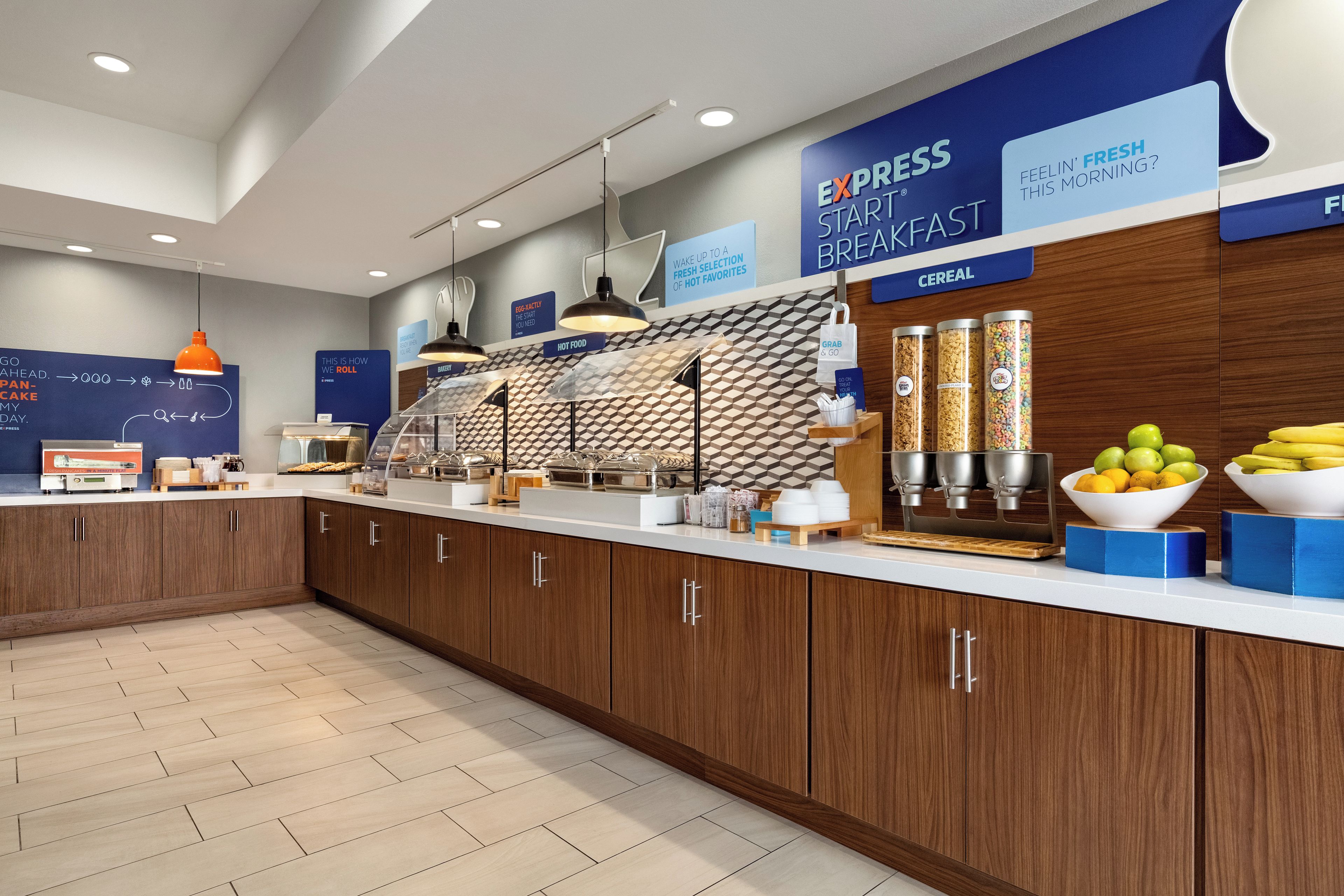 Un área de desayuno moderna con mostrador, estación de alimentos calientes, dispensadores de cereales y un cartel que indica las opciones de desayuno.