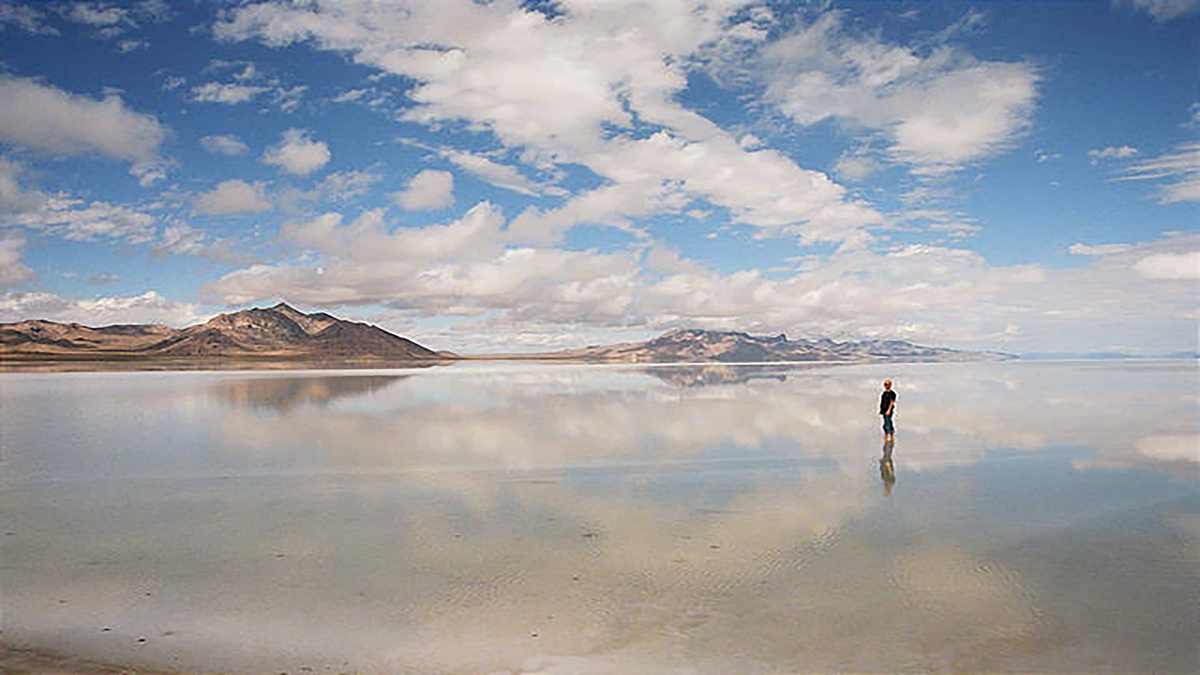 Eine Person steht auf einer spiegelnden Wasseroberfläche, im Hintergrund Berge.