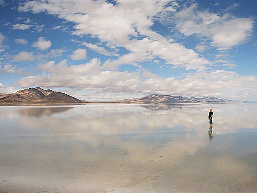 Eine Person steht auf einer spiegelnden Wasseroberfläche, im Hintergrund Berge.