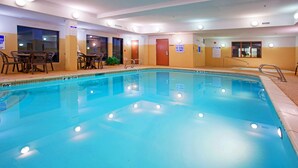 An indoor swimming pool with clear water, a shallow water sign, and a table with chairs on the side.
