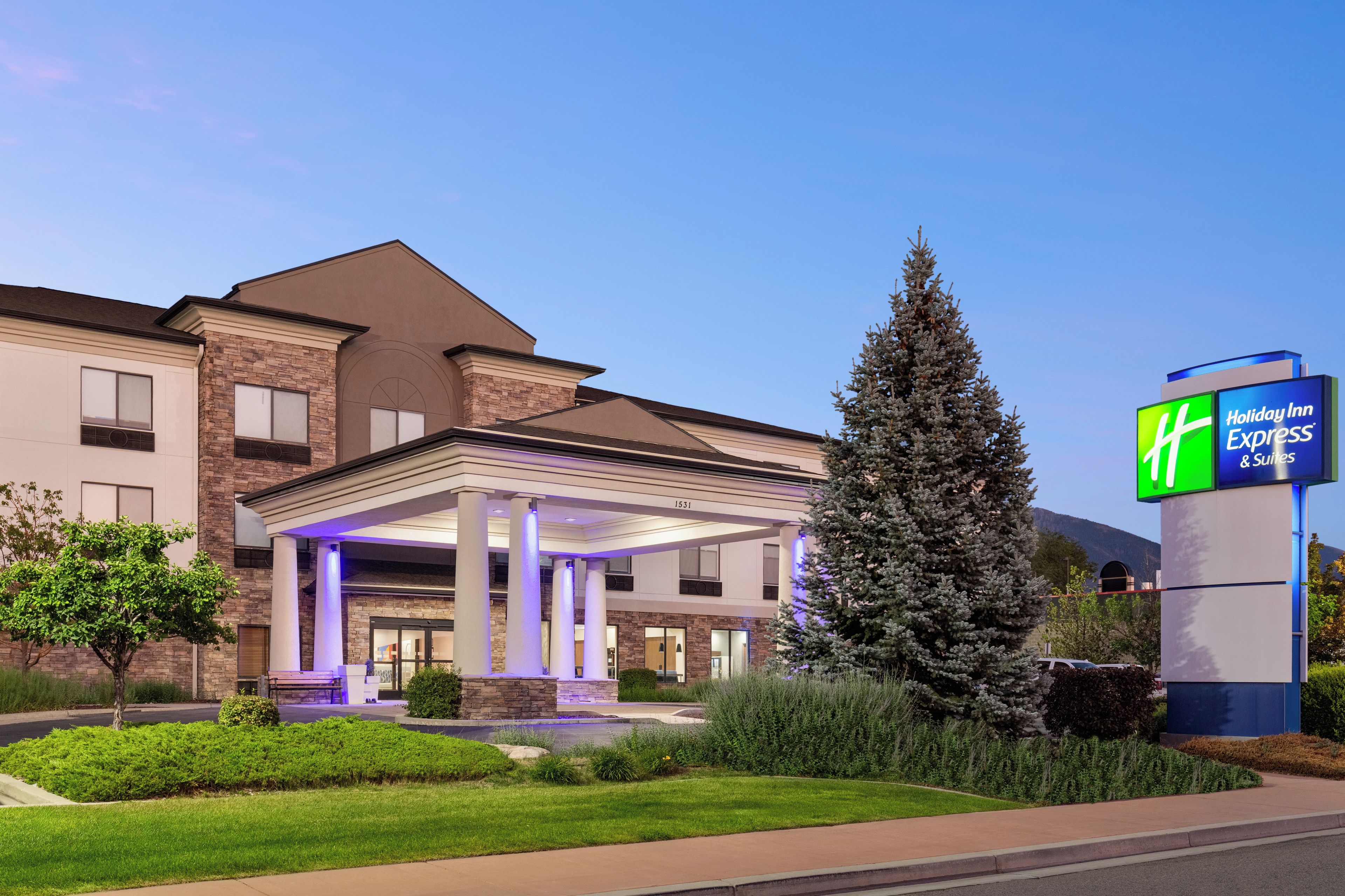 A Holiday Inn Express & Suites building with a covered outdoor area, a large evergreen tree, and a well-maintained lawn.