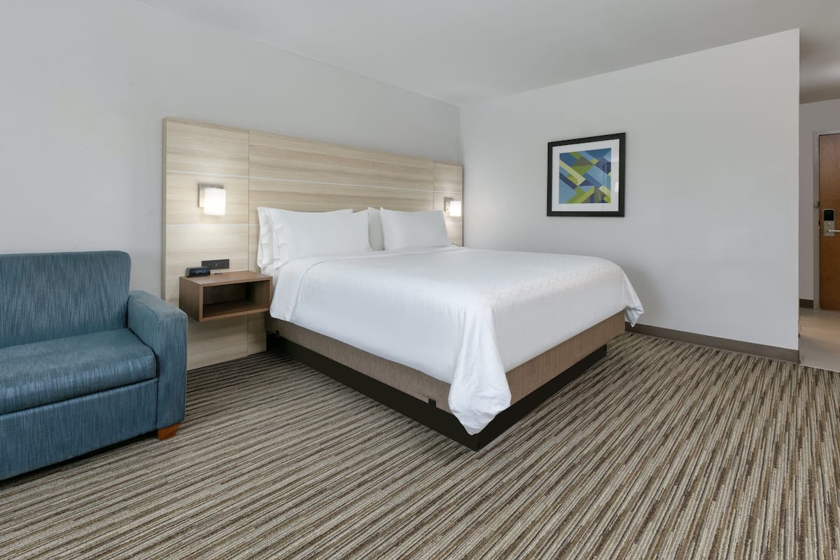 Quarto de hotel com uma cama, uma poltrona azul, uma mesa de cabeceira e um quadro na parede.