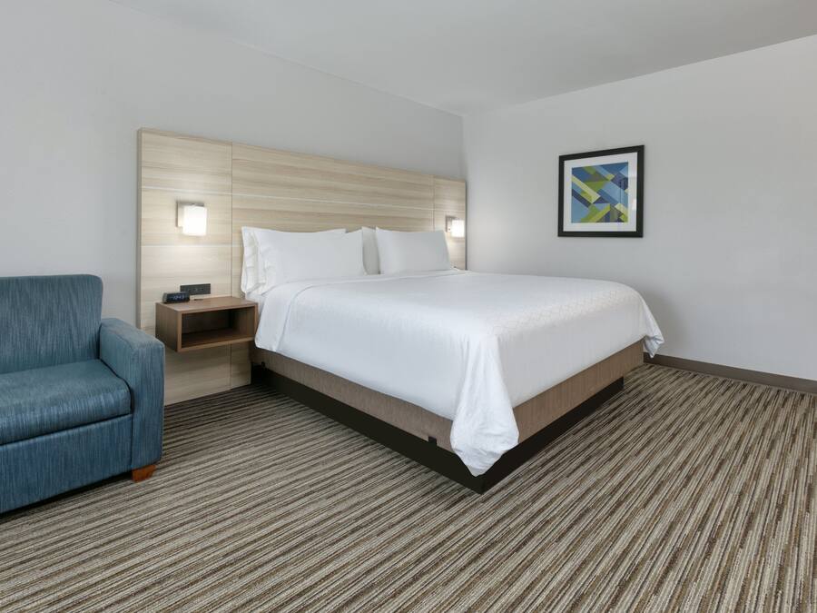 Ein Hotelzimmer mit einem Bett, einem blauen Sessel, einem Nachttisch und einem gerahmten Bild an der Wand.