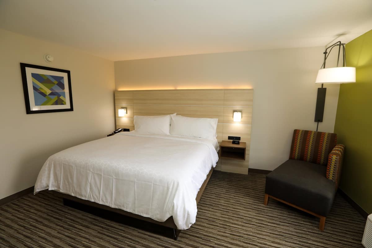 Quarto de hotel com uma cama grande, mesas de cabeceira, uma cadeira e uma luminária fixada na parede.