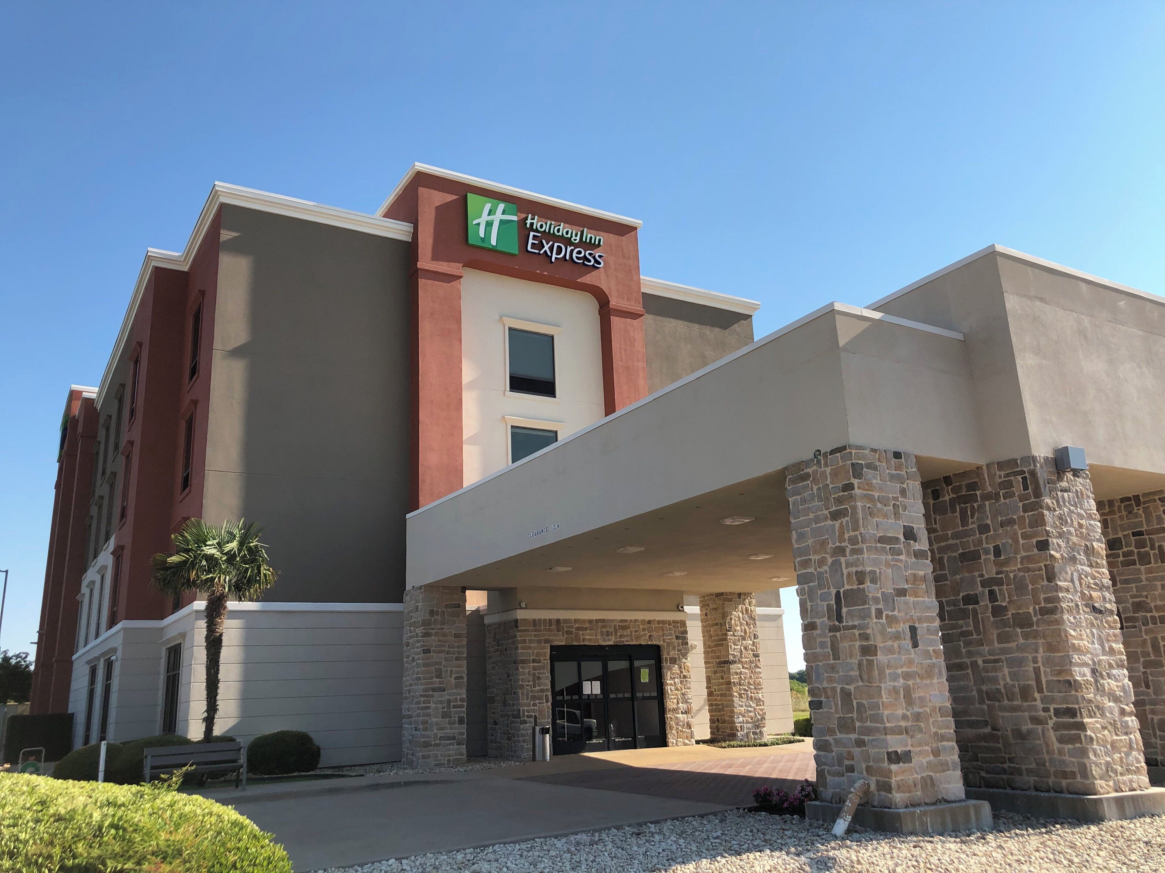 Un bâtiment de l’hôtel Holiday Inn Express avec une façade en pierre, une entrée couverte et un palmier au premier plan.