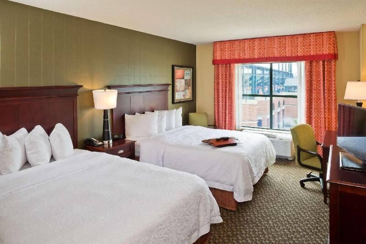 Une chambre d’hôtel avec deux lits, un bureau, une chaise, une lampe et une fenêtre avec des rideaux.