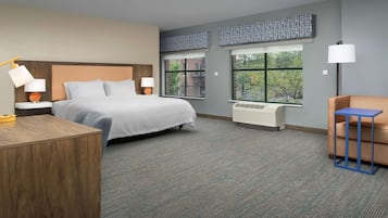 Una habitación de hotel con una cama grande, mesitas de noche con lámparas, un escritorio, una silla y ventanas amplias con vistas a árboles.