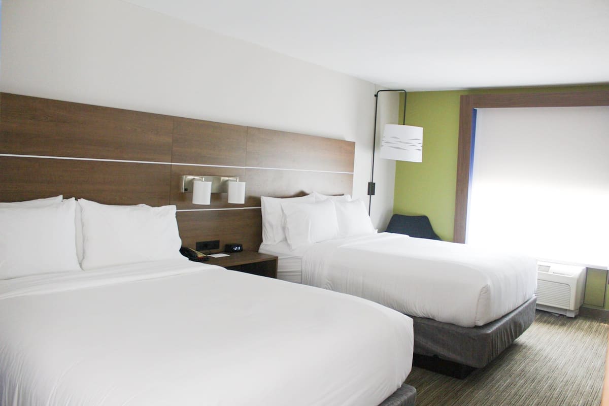 Une chambre d’hôtel avec deux lits, une table de chevet avec une lampe, une horloge et une fenêtre donnant sur un paysage agréable.
