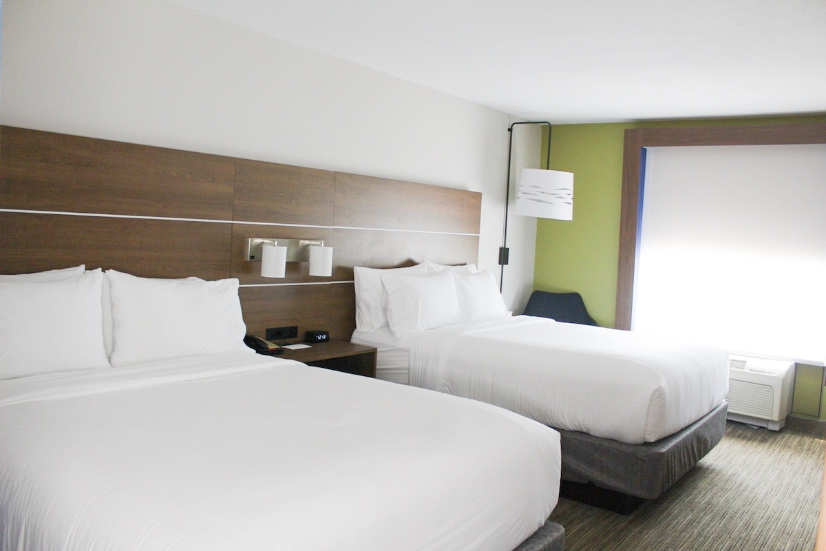 A hotel room with two beds, a bedside table with a lamp, a clock, and a window with a view.
