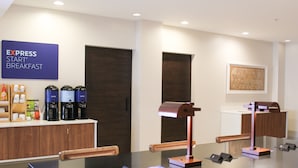En modern hotellfrukostsal med en skylt som lyder ”EXPRESS START BREAKFAST”, kaffemaskiner, ett skrivbord med en lampa och ett inramat konstverk på väggen.