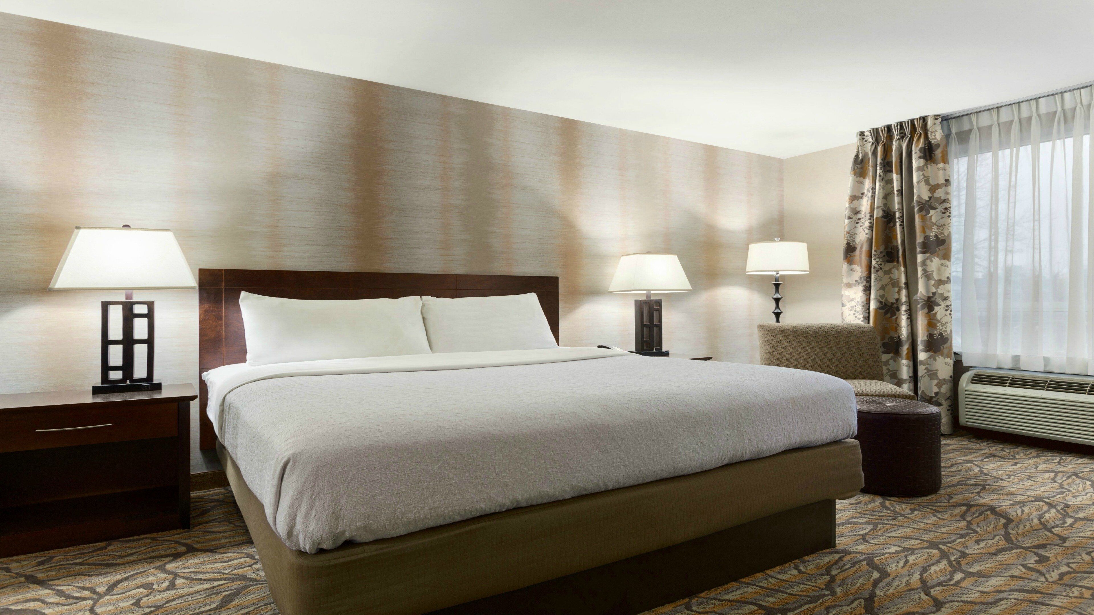 A hotel room with a large bed, two bedside tables with lamps, a chair, a small table, and a window with curtains.