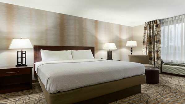 A hotel room with a large bed, two bedside tables with lamps, a chair, a small table, and a window with curtains.