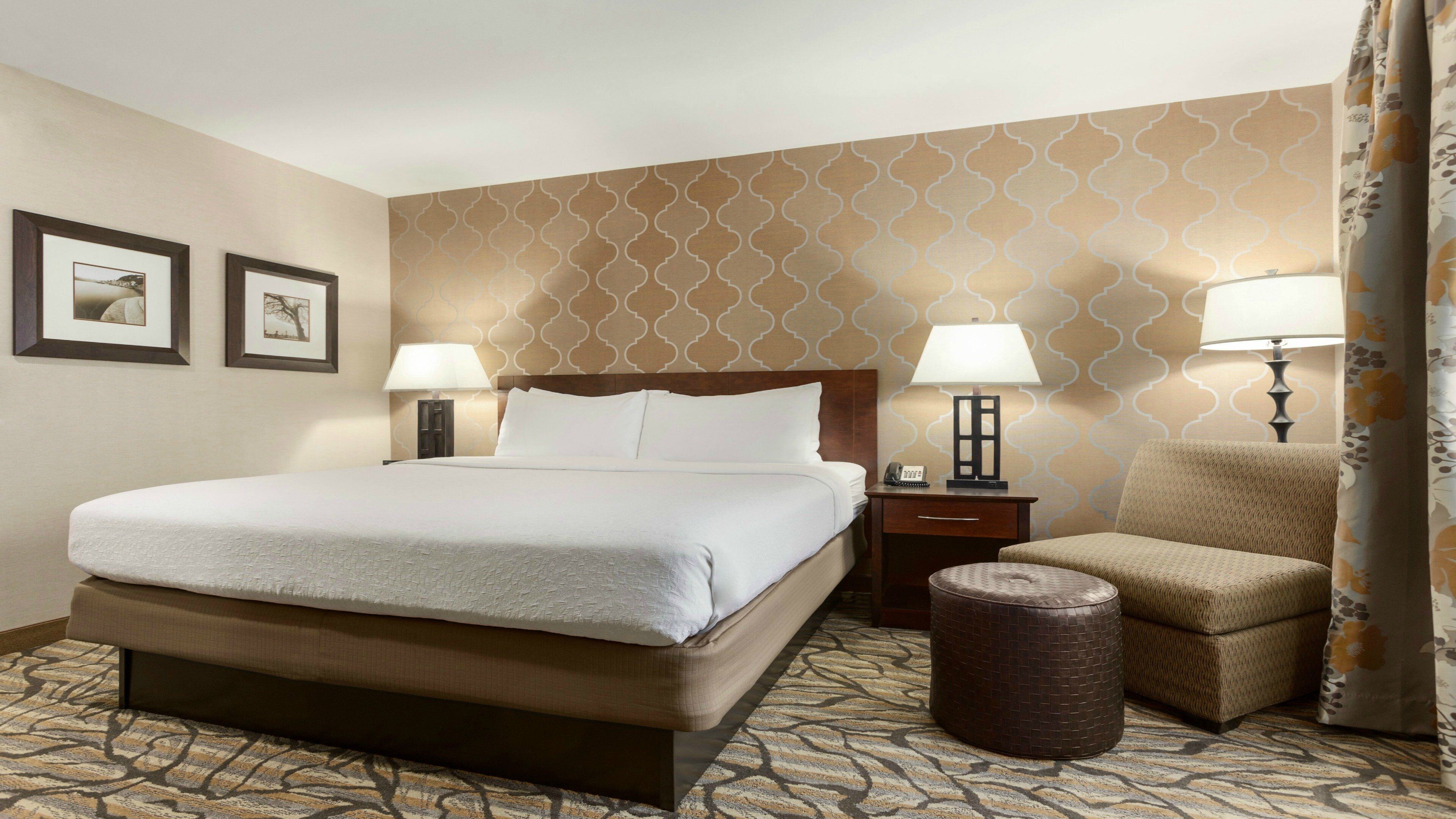 A hotel room with a large bed, two bedside tables, a chair, a small ottoman, and patterned wallpaper.
