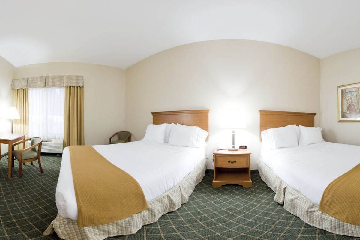 A hotel room with two beds, a desk, a chair, and a nightstand with a lamp.