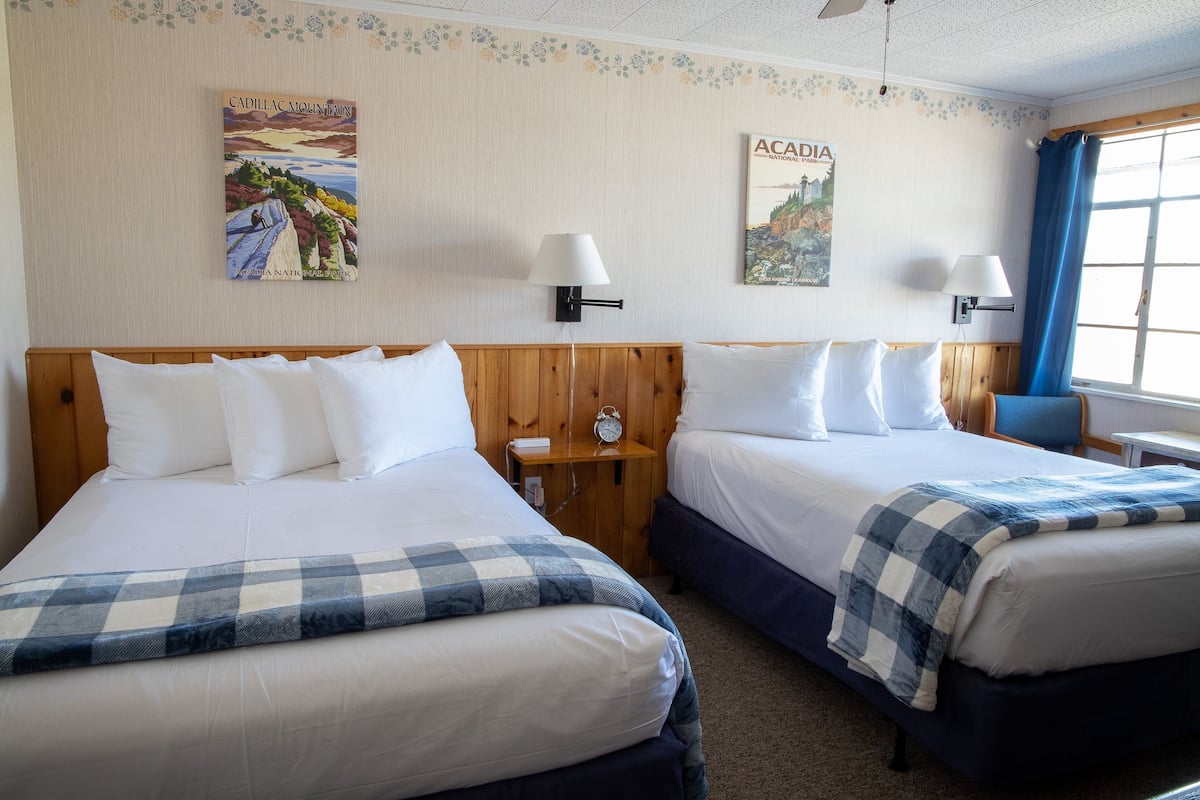 A hotel room with two beds, a wooden headboard, wall-mounted lamps, and two posters.