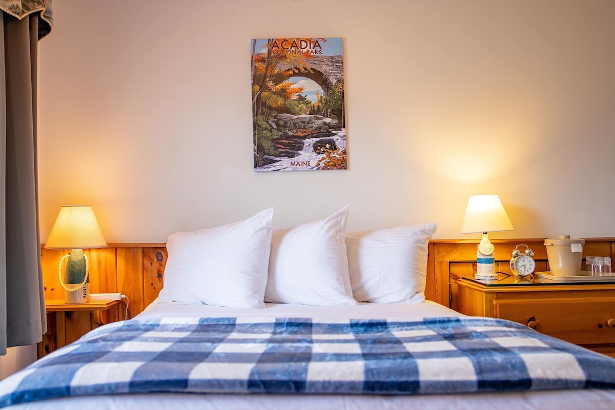 A hotel room with a bed, two bedside tables with lamps, a poster of Acadia National Park, and a wooden nightstand with a clock and a bottle.